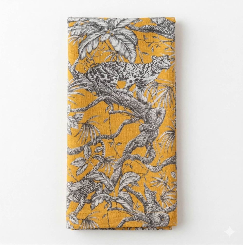A close-up of the Leonard Mustard Linen tablecloth showcases detailed black-and-white leopard jungle motifs with tree branches and leaves, elegantly draping over the table's edge.