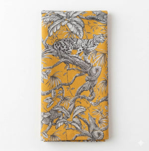 The Leonard Mustard Linen showcases an intricate black and white leopard jungle motif, with leopards lounging and climbing among leaves on a folded mustard linen fabric with a yellow background.