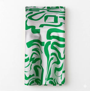 The Mila Linen napkin, folded to display its abstract green and white pattern of curving lines, sits on a plain light background—ideal for bringing modern elegance to contemporary event décor.